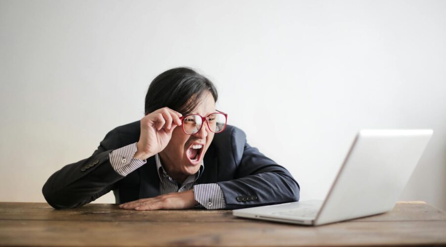 Modern Asian man in jacket and glasses looking at laptop and screaming with mouth wide opened on white background