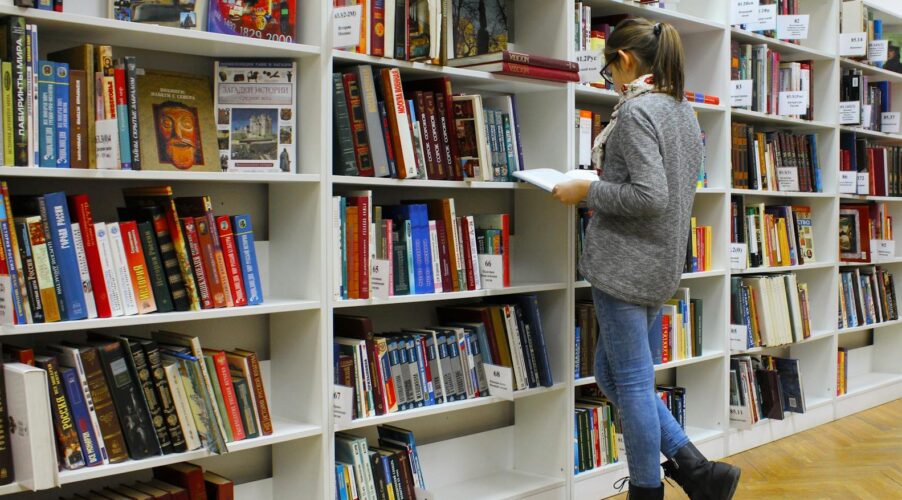 A young woman stands reading a book in a well-stocked library.