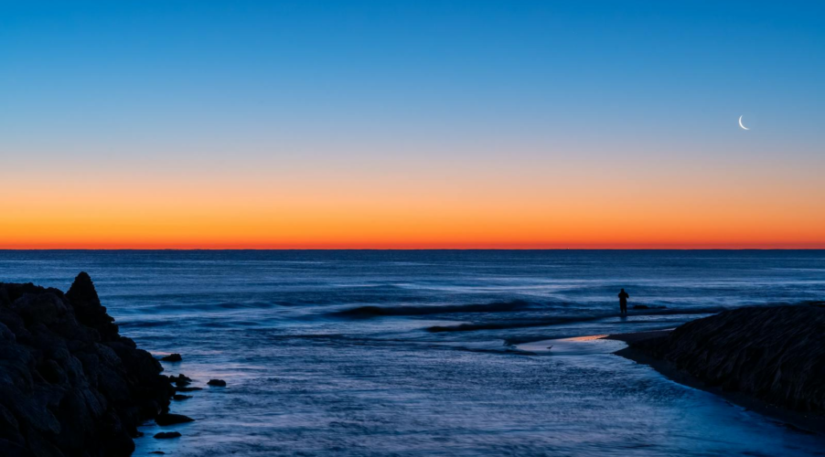 A tranquil twilight scene of a beach with a crescent moon and silhouette of a fisherman in Valencia, Spain.