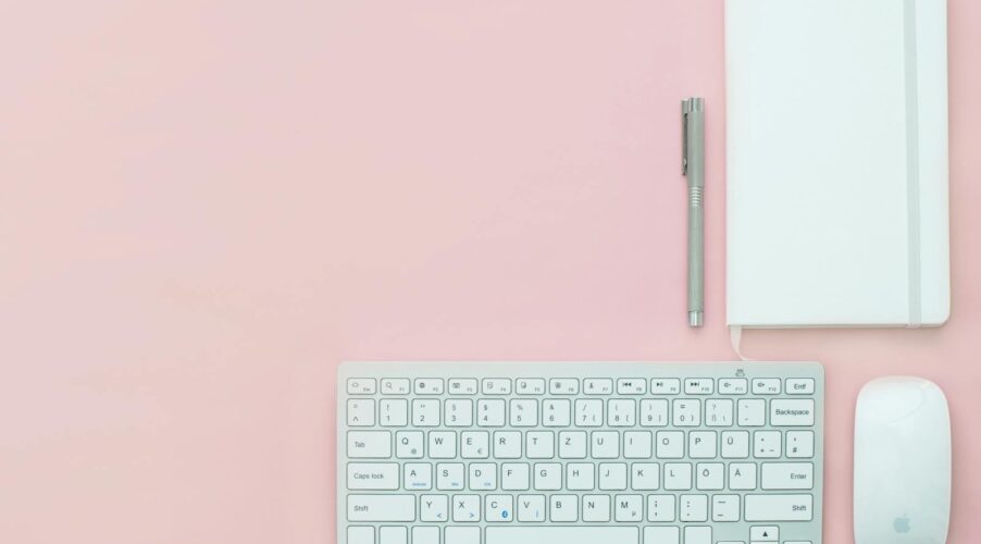 Flat lay of a minimalist workspace featuring a keyboard, notebook, and pen on a pink surface.