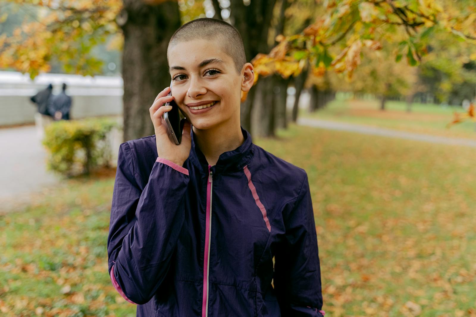 Cheerful woman with shaved head talking on phone in a fall park setting, enjoying an outdoor walk.