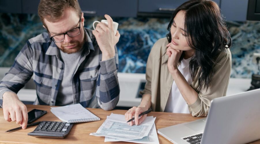 A couple reviews bills and documents on a laptop while discussing household finances.
