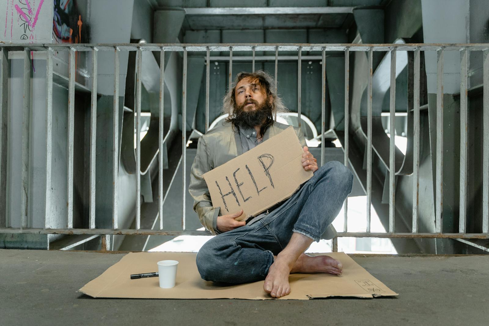 A homeless man sits under a bridge with a sign reading 'HELP', highlighting poverty and homelessness.