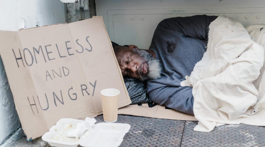 Elderly homeless man sleeping on the street with a sign reading 'Homeless and Hungry'.